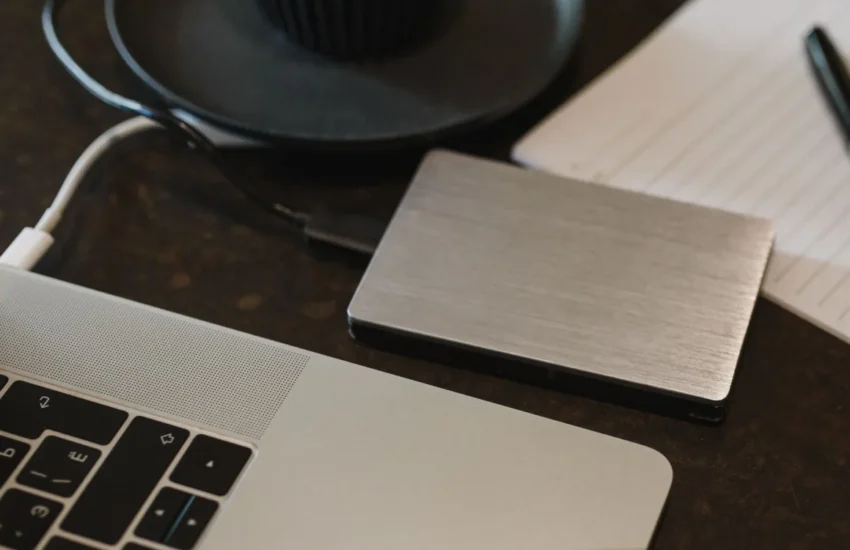Zewnętrzny dysk w metalowej obudowie leży obok laptopa, w tle notatnik i pióro
