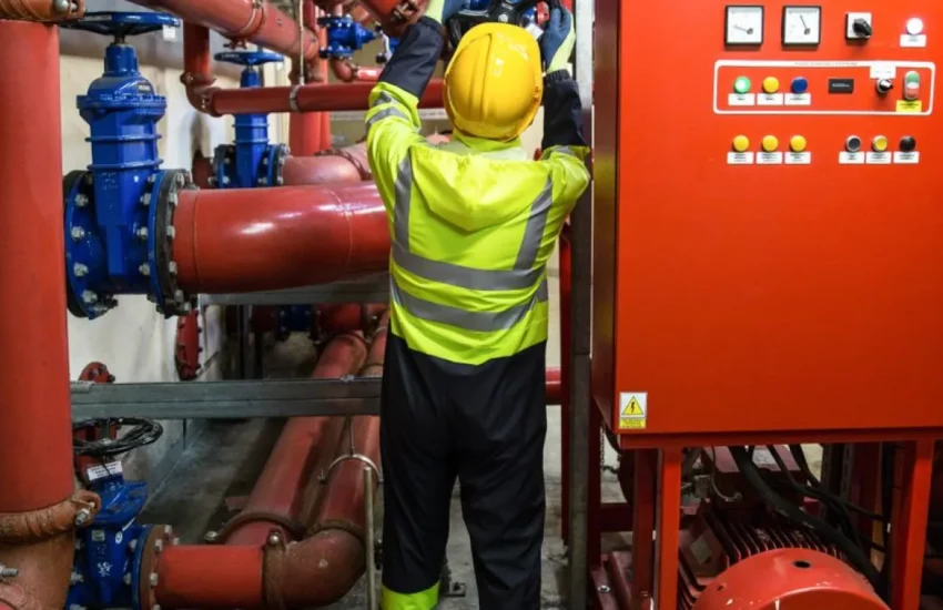 Technik w kasku i odblaskowej kurtce przy czerwonych rurociągach i szafie sterowniczej